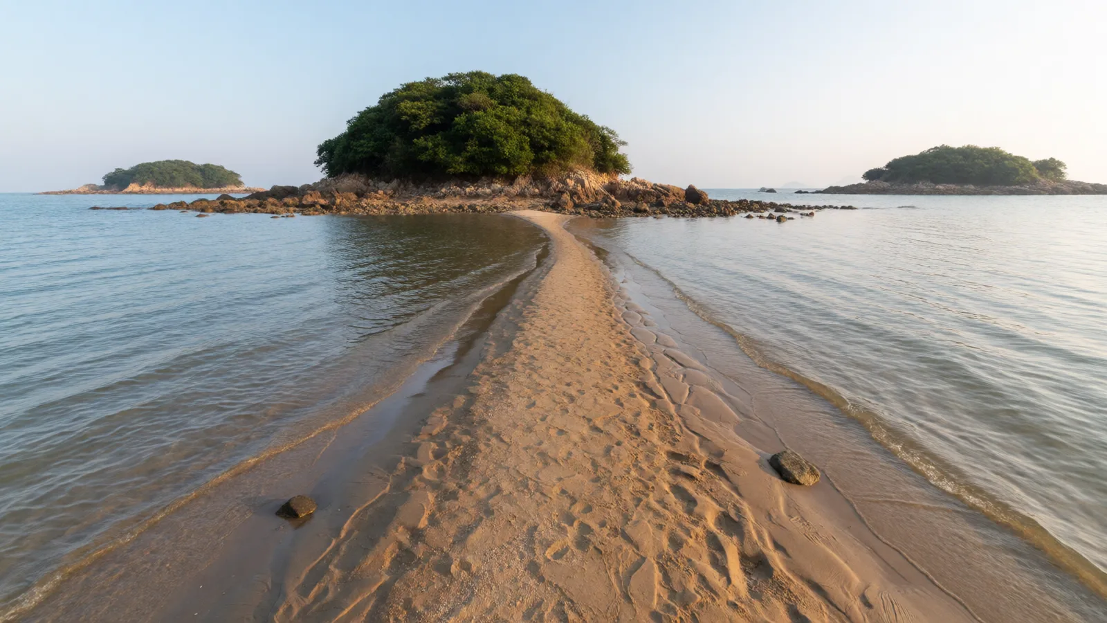 退潮的早晨，一條細長的沙洲從岸邊延伸到有岩石的綠色小島，兩側是平靜的淺海（AI生成圖片）