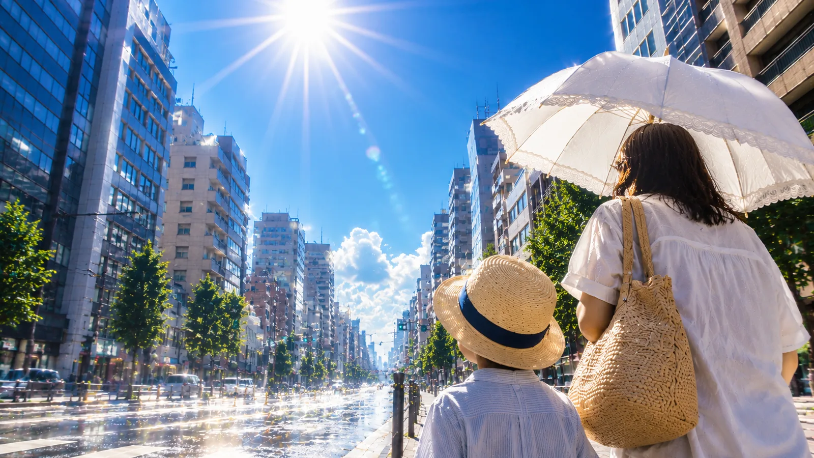 真夏の強い日差しが照りつける日本の街を、日傘と帽子で歩く親子のイメージ
